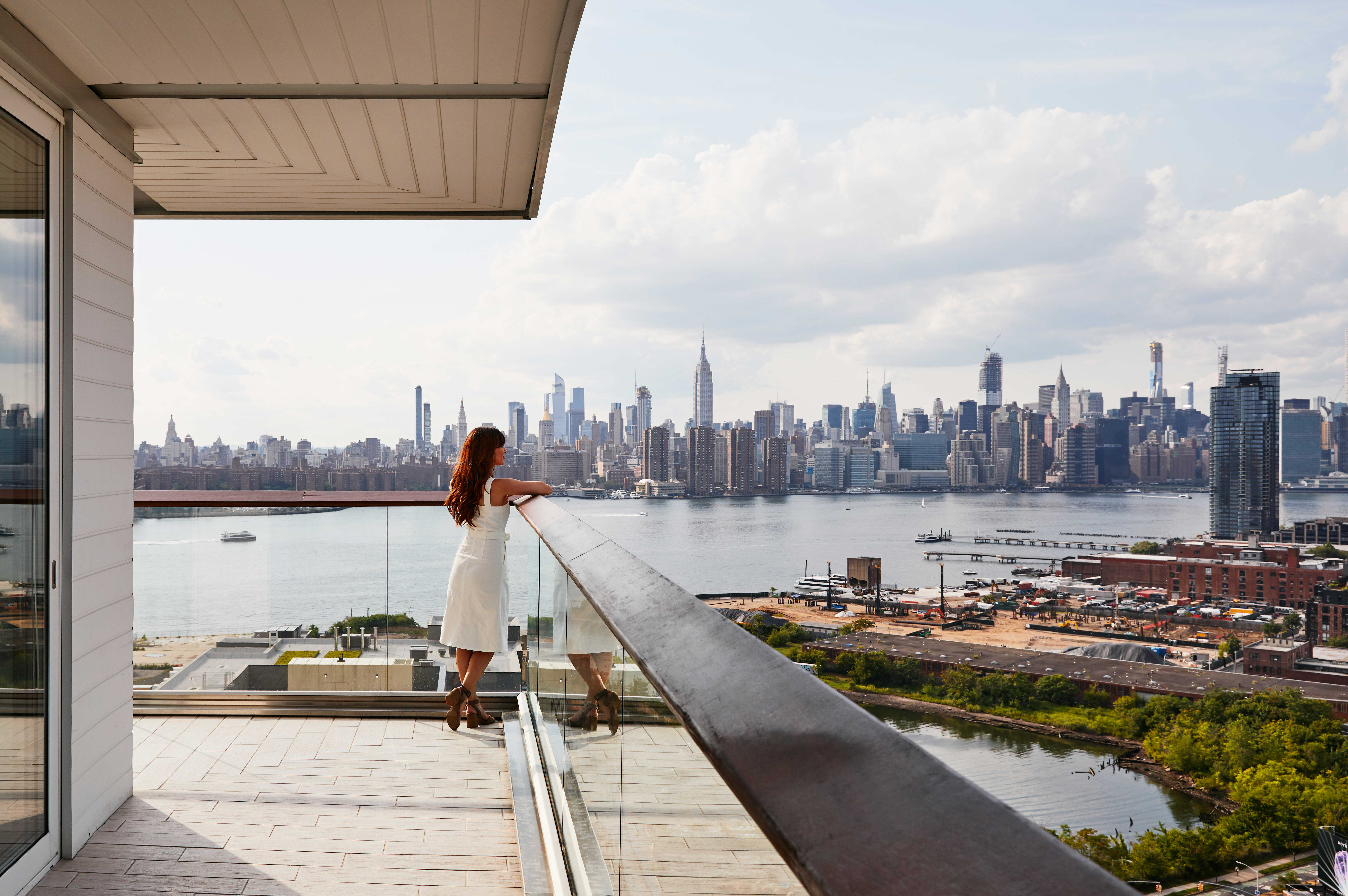 A woman stands on a balcony with a glass railing, looking out at the city skyline across the water.